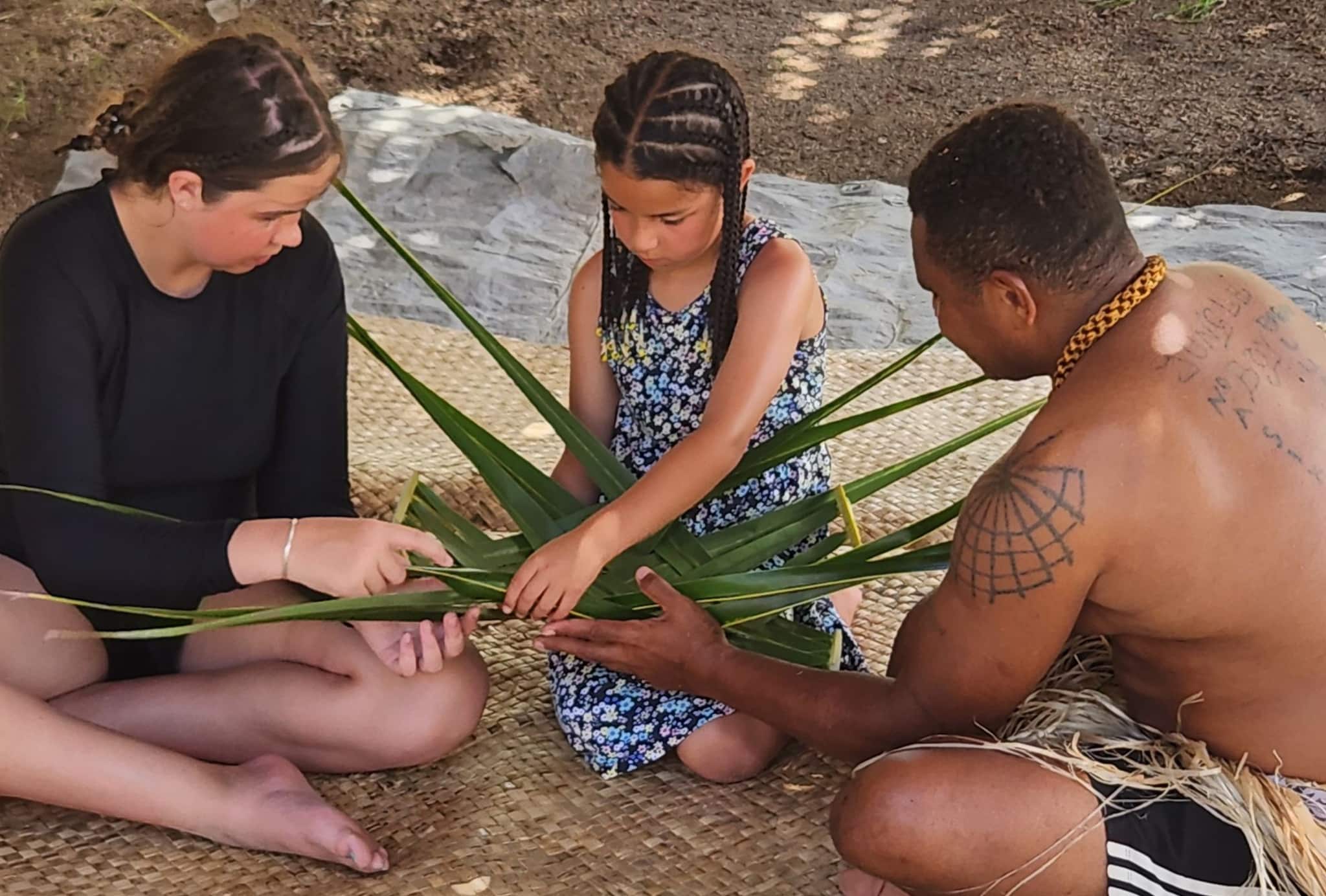 Two girls weaving a flax basket with the help of a man.